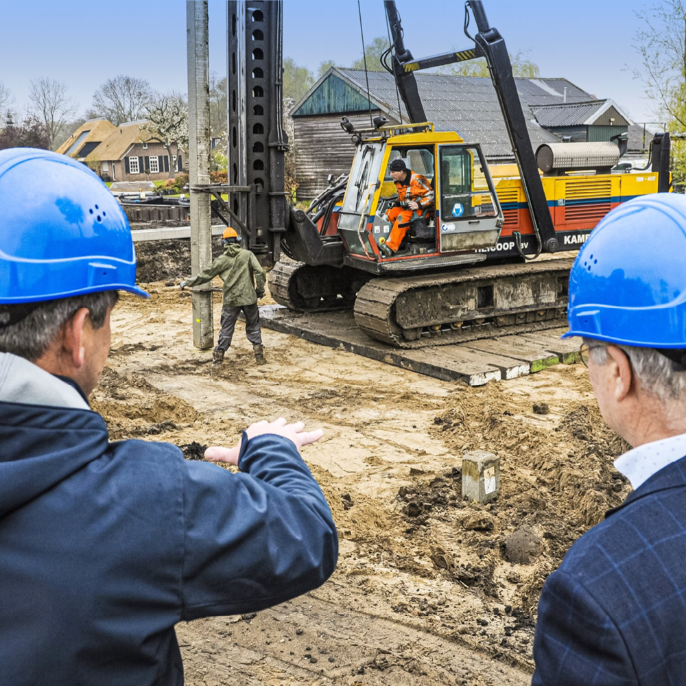 Mensen met blauwe helmen bekijken een bulldozer die op een bouwplaats een betonnen paal in de grond plaatst.