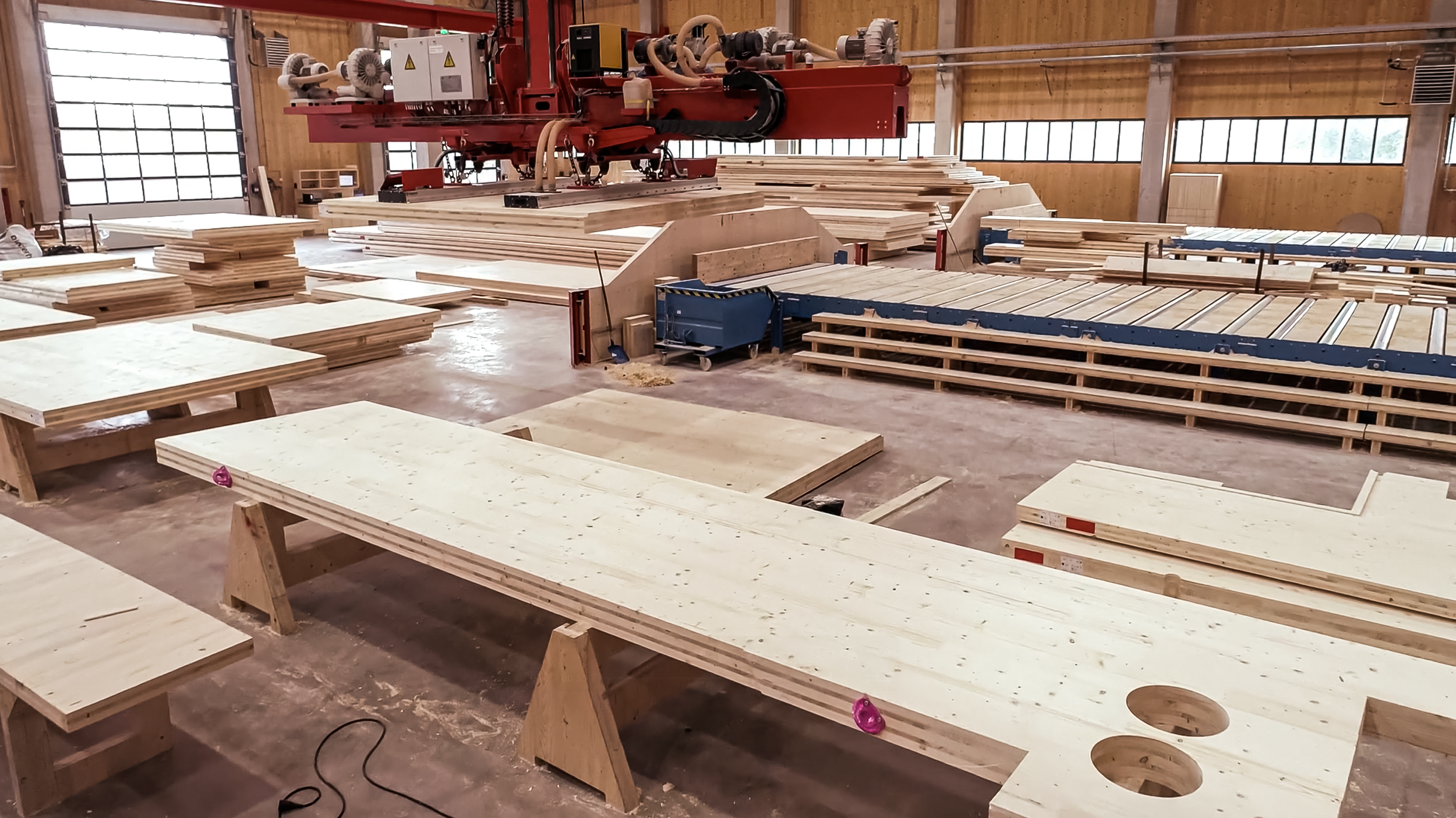 Houten werkplaats met grote zaagmachine, meerdere werkbanken, planken en houtbewerking apparatuur.