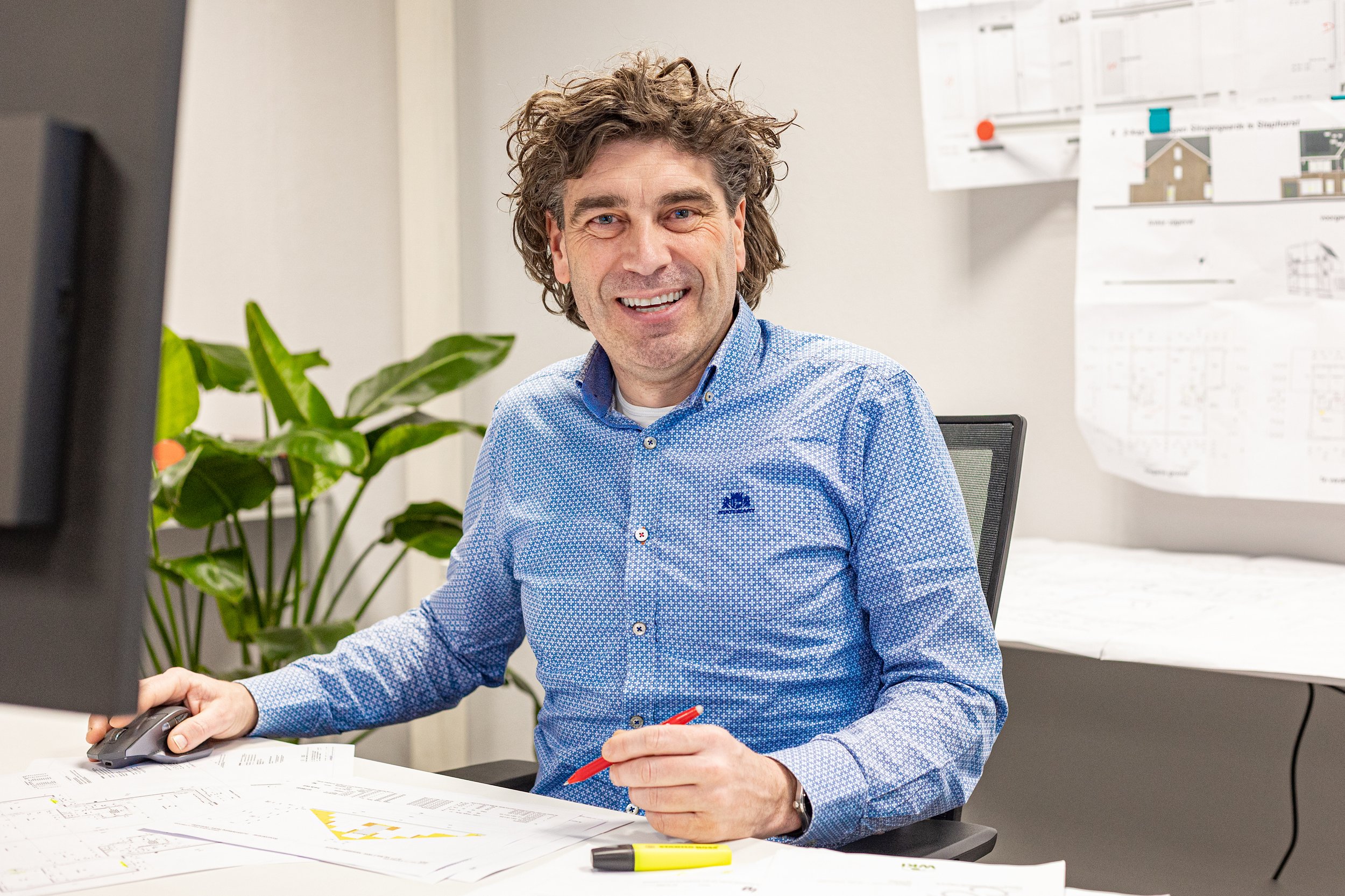 Man op kantoor met bouwplannen, lachend, met computer en groene plant op de achtergrond.