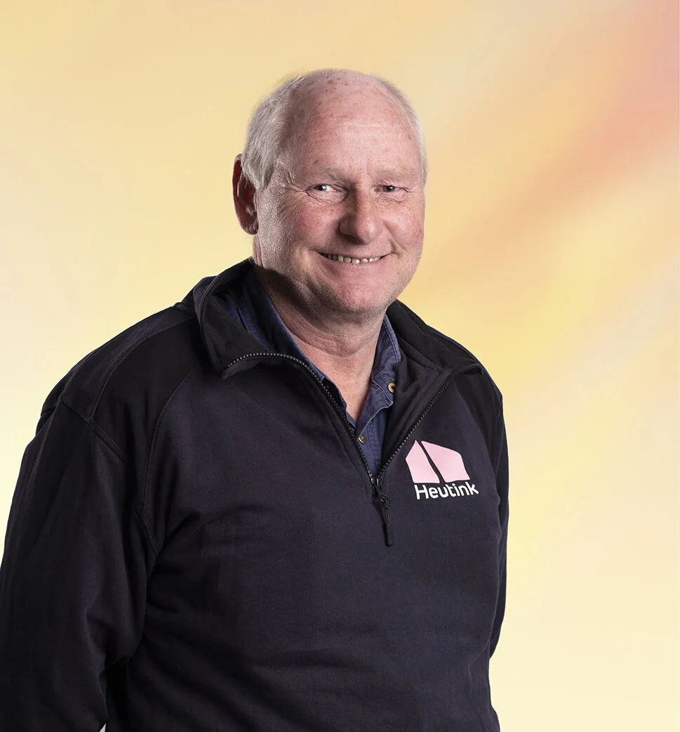 Een oudere man met een glimlach, draag een zwart jasje met een logo en achtergrond die een zachte gradient van geel, oranje en roze heeft.