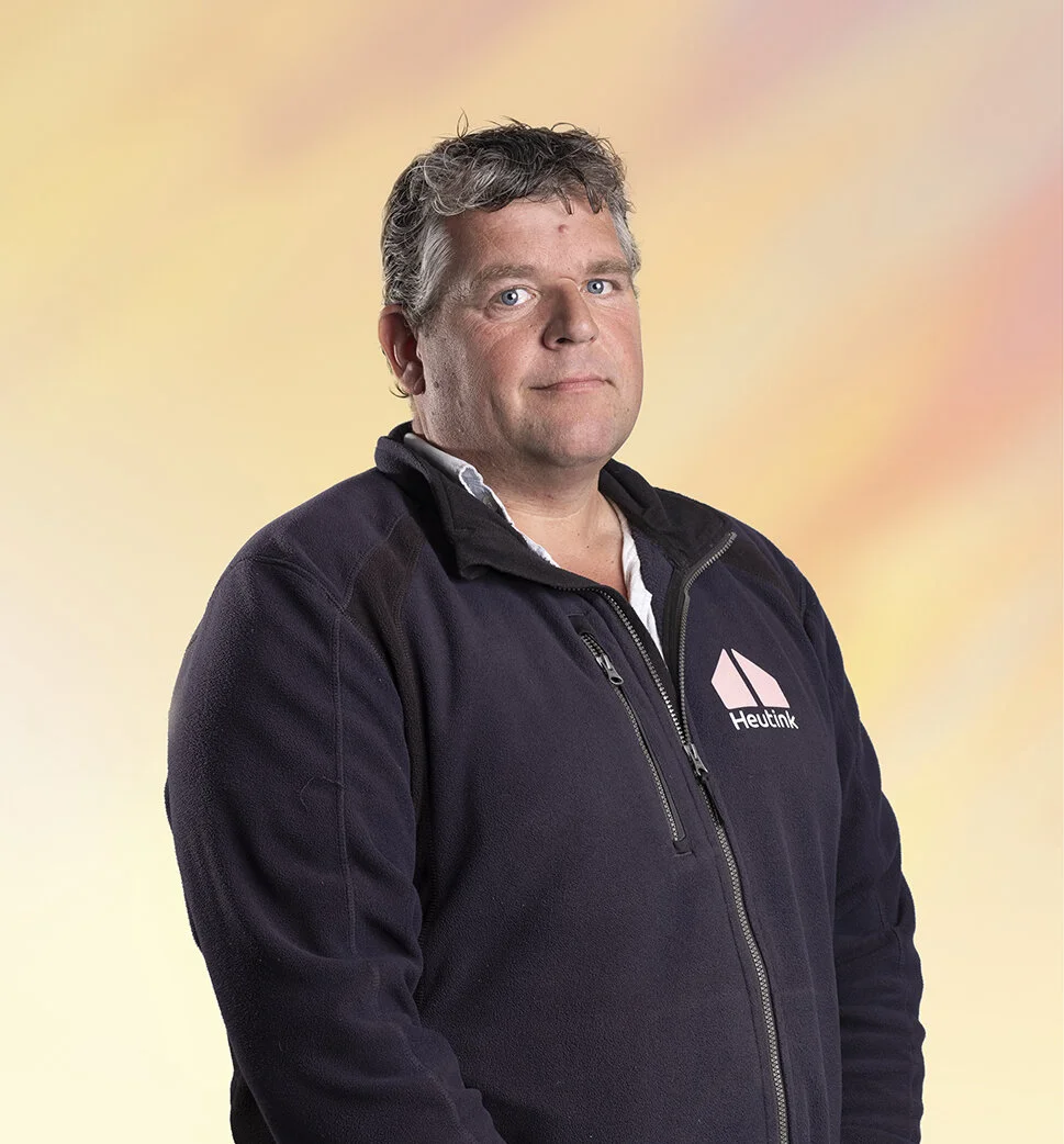 Man met grijs haar en blauwe ogen, draagt een zwarte hoodie met het logo van Heutink, voor een pastelkleurige achtergrond.
