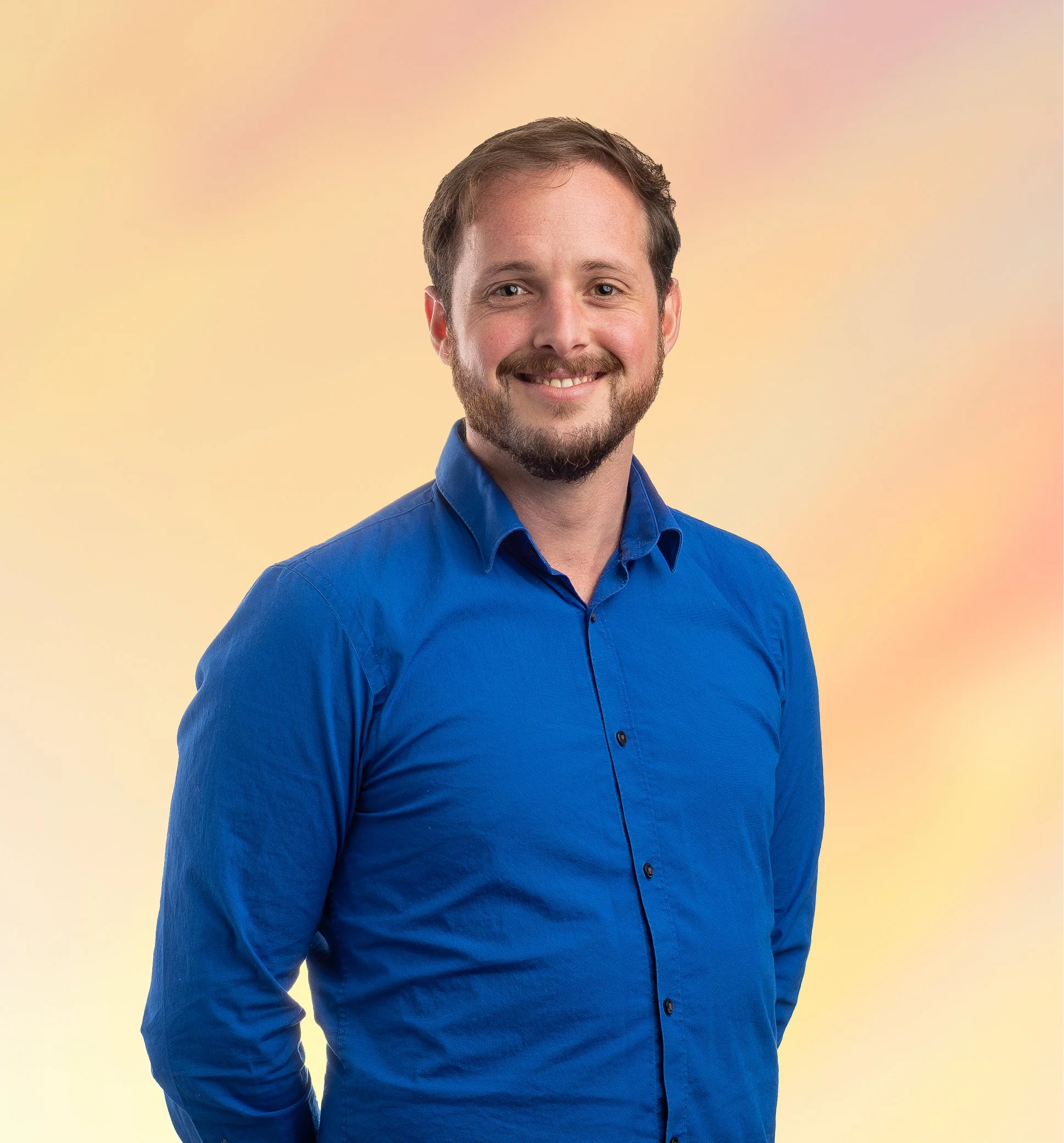 Portret van een man met een baard en kort bruin haar, lachend en gekleed in een blauw overhemd, staand voor een gele en roze achtergrond.
