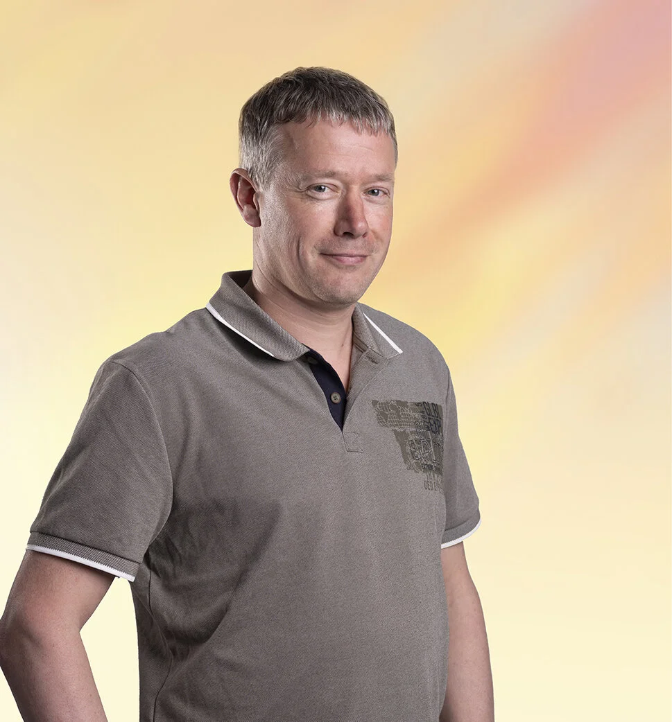 Een man met kort bruin haar draagt een grijze polo met een opdruk aan de linkerborst en staat voor een pastelkleurige gradient achtergrond.