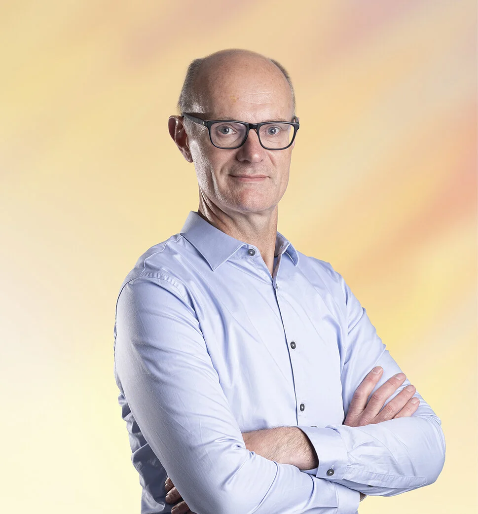 Een oudere man met bril, gekleed in een lichtblauw overhemd, staat met versch crossed armen voor een kleurrijke, zachte achtergrond.