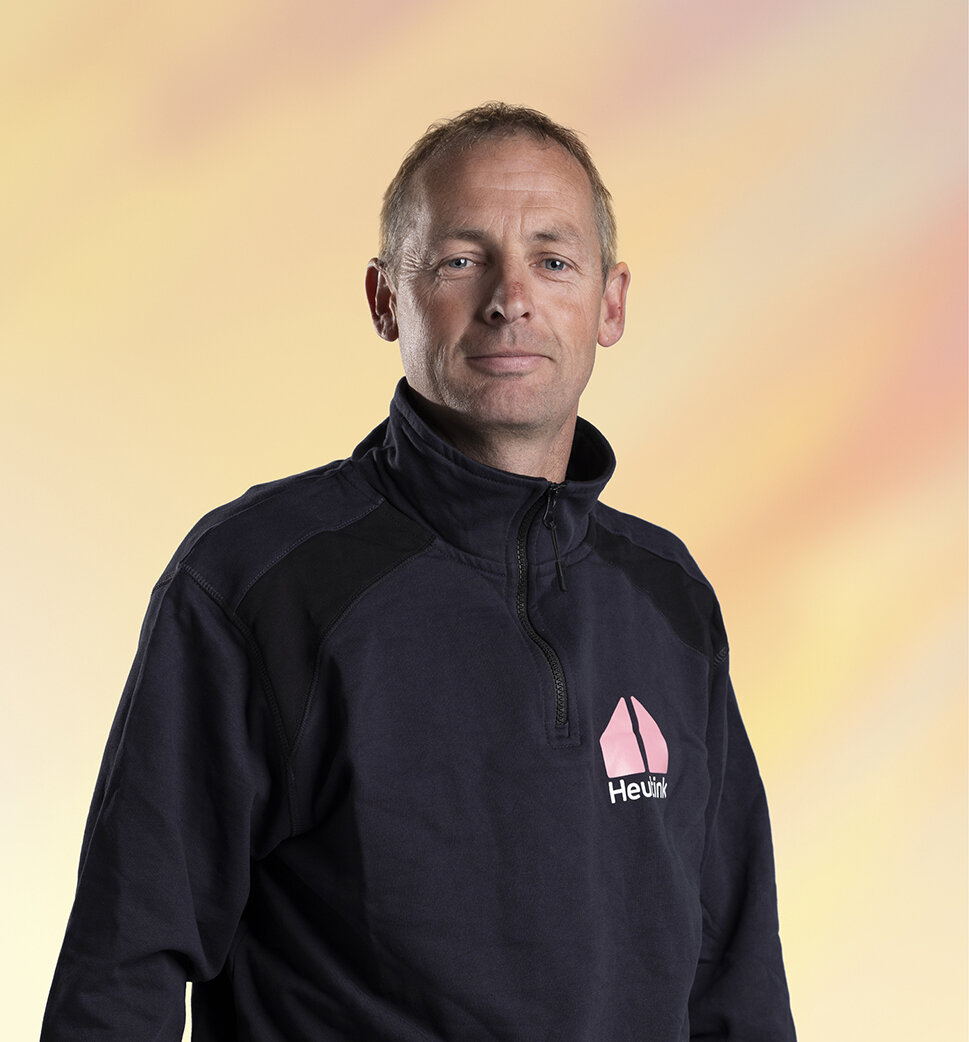 Man draagt een donkerblauwe jas met het logo van Heuink, staat voor een pastelkleurige achtergrond.