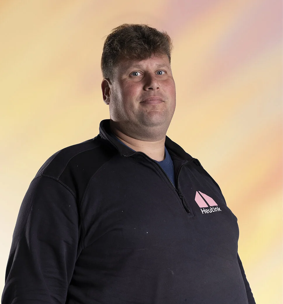 Portret van een man met kort bruin haar, dragen een zwarte jas met een logo op de borst tegen een zachte pastelgekleurde achtergrond.