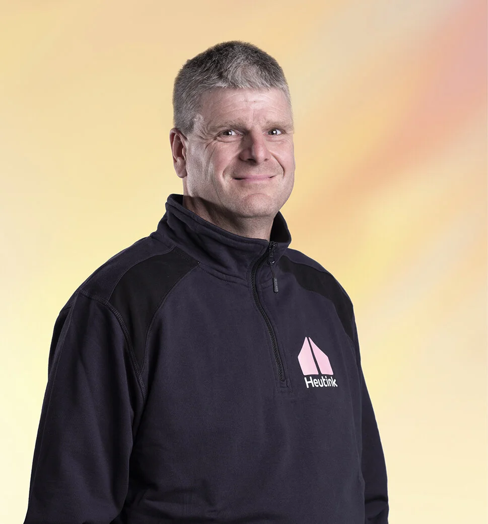 Man draagt een zwart vest met een Heutink logo, staat voor een pastelkleurige achtergrond, glimlacht licht.