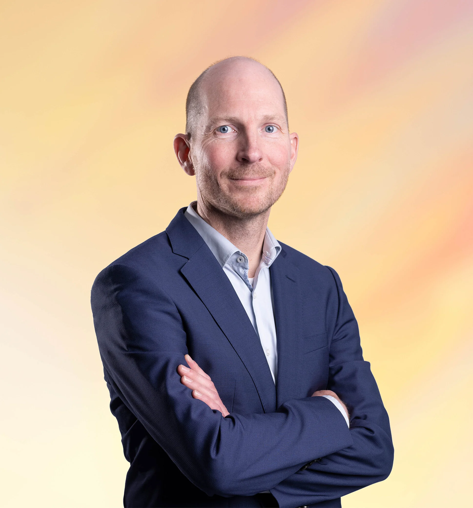 Man in een blauwe blazer met gekruiste armen, glimlachend, tegen een pastelkleurige achtergrond.