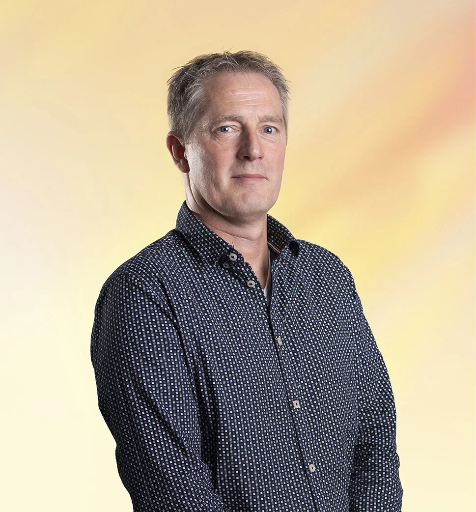 Portret van een volwassen man met kort, grijs haar, gekleed in een donkerblauw overhemd met kleine witte patronen, staand voor een zachte gele en roze achtergrond.