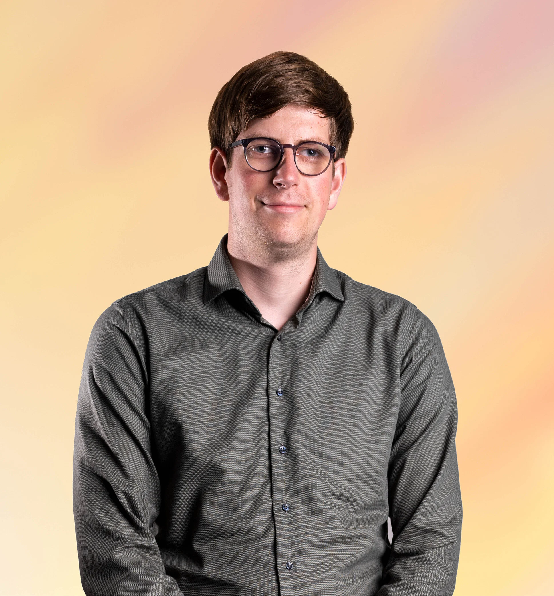 Portret van een jonge man met bruin haar, brilmontuur, in een grijze shirt met knopen, lachend, achtergrond met pastelkleuren.
