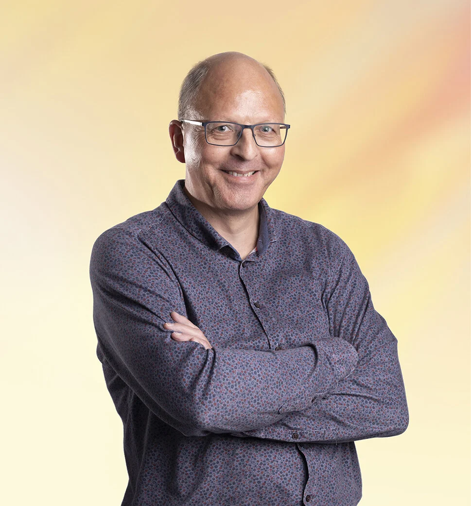Man met bril, glimlach en gekruiste armen, gekleed in een blouse met een patroon, voor een kleurverloop achtergrond.