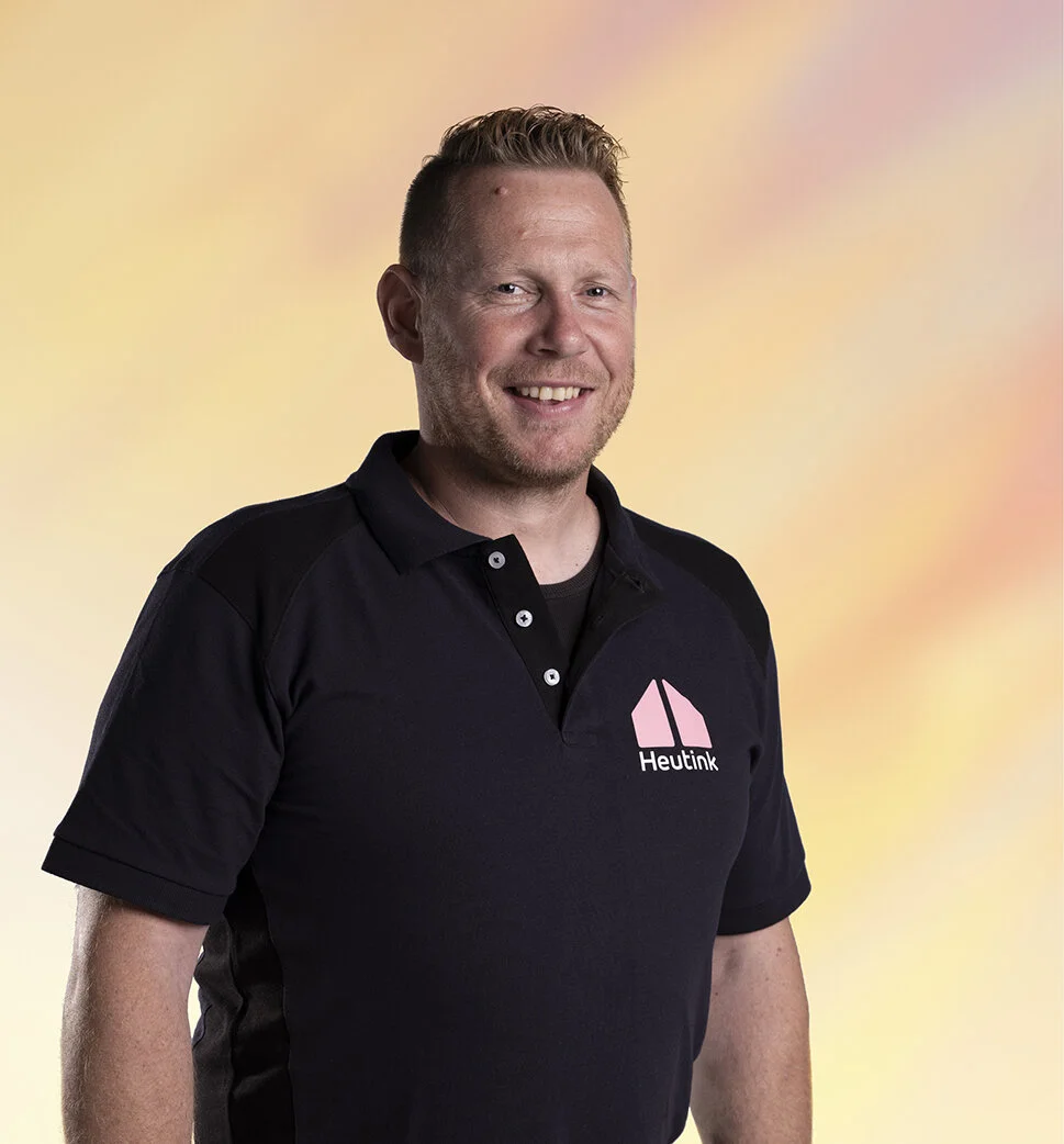 Man in zwart bedrijfspolo met logo Heutink, glimlachend, kleurrijke achtergrond.