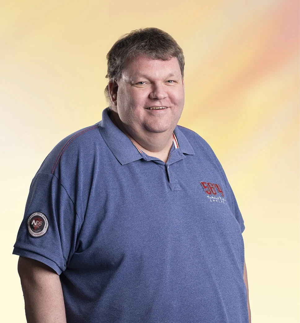Portret van een lachende man met kort bruin haar in een blauwe polo met logo's, staand voor een gele en roze achtergrond.