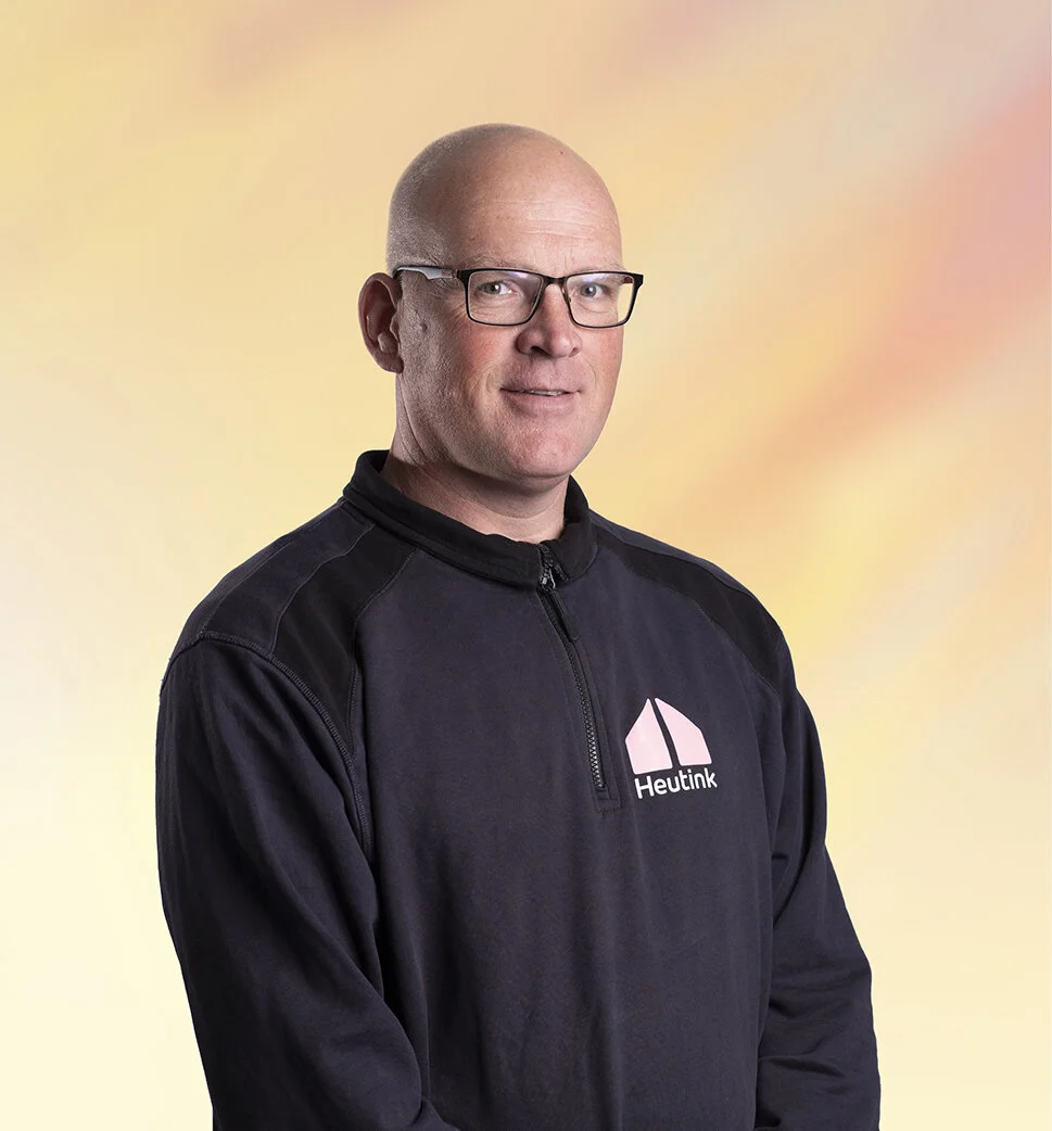 Een man met een kale kop draagt een donkere sportjack met een logo en tekst 'Heutink' op de voorkant, staat voor een geschilderde achtergrond in pastelkleuren.