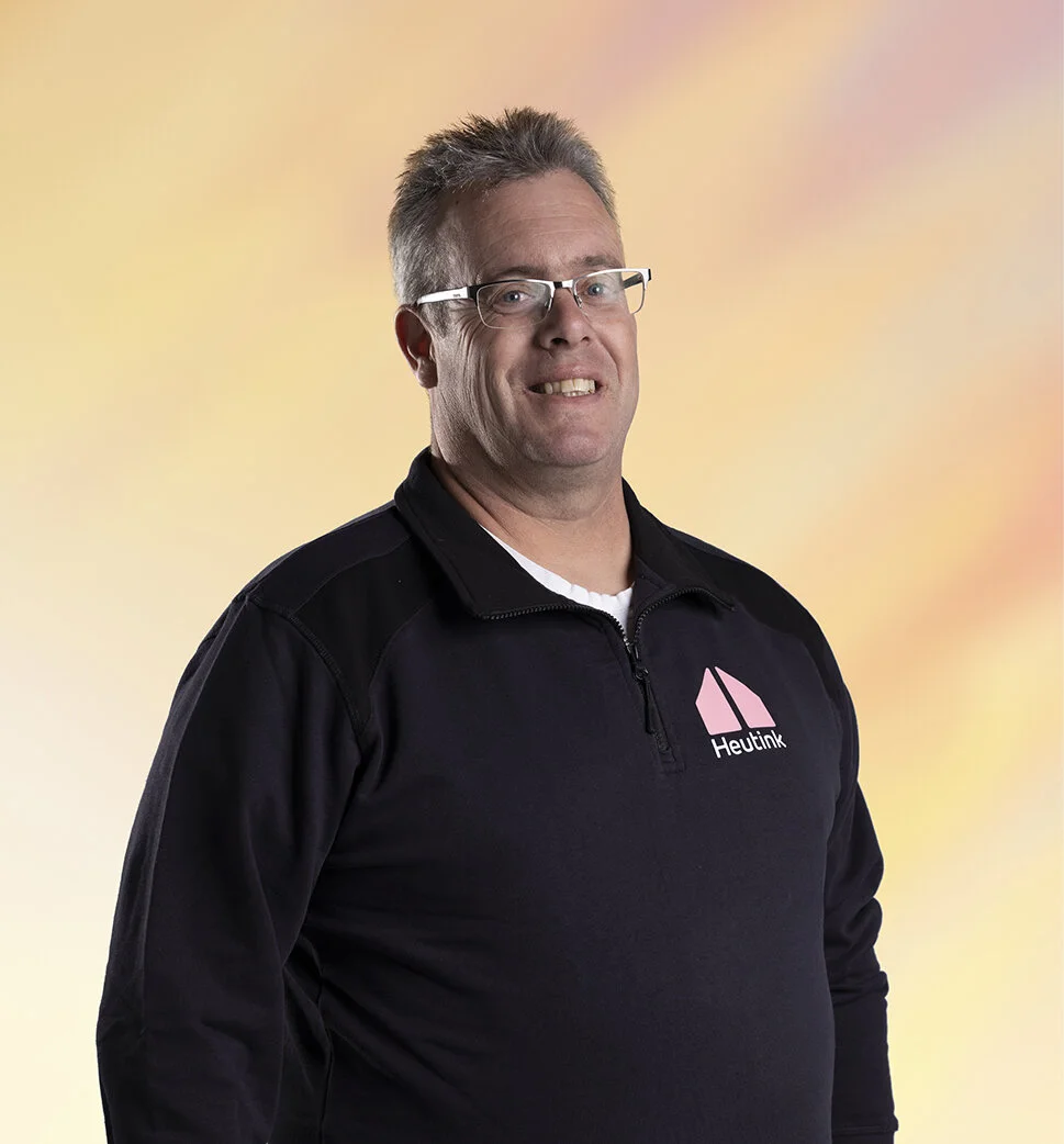 Een man met kort, grijs haar, bril en een zwart arc jacket met een pink logo en de tekst 'Heutink', poseert voor een pastelkleurige achtergrond.