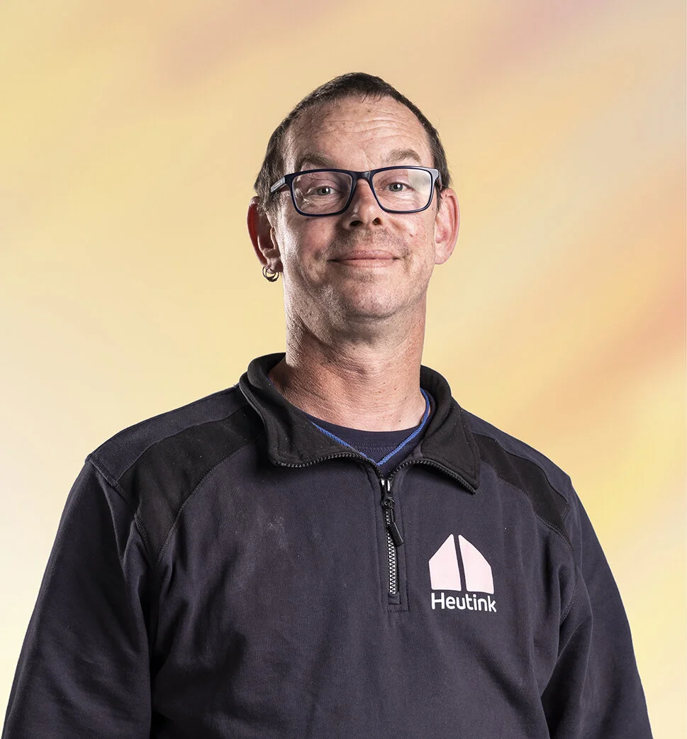 Man met kort haar, brillen en een donkere trainer met het logo 'Heutink' op de voorkant, lachend voor een kleurrijke achtergrond.