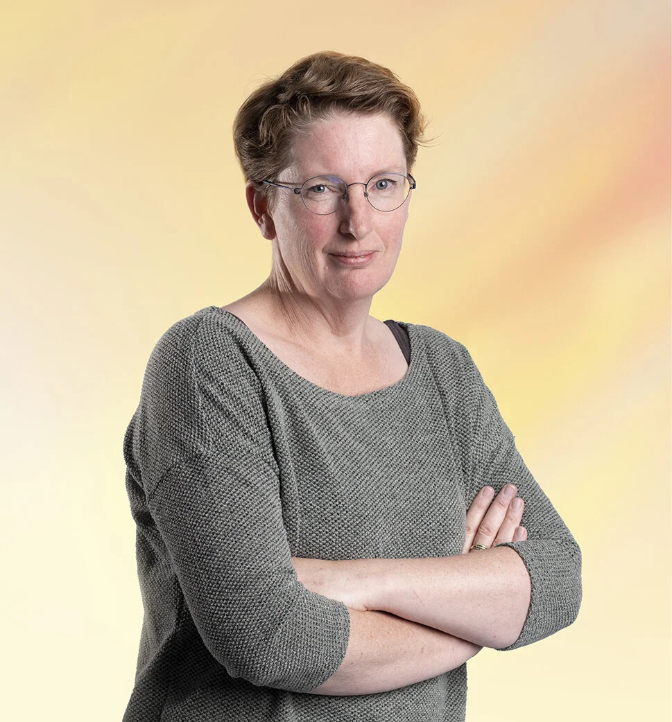 Vrouw met kort bruin haar, bril en grijze trui staat met gekruiste armen voor een pastelkleuren achtergrond.