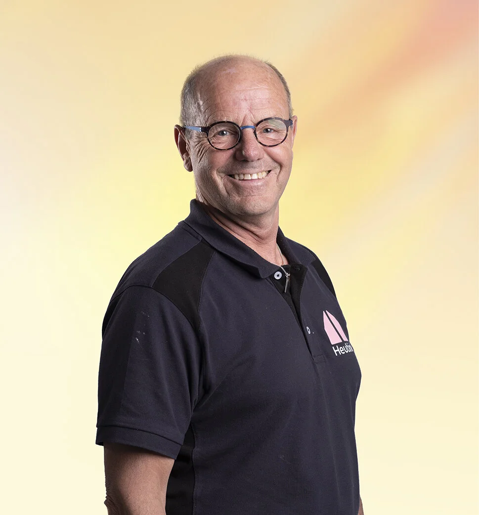 Een glimlachende man met bril, een zwart polo met een logo, staand voor een zachte, gele en roze achtergrond.