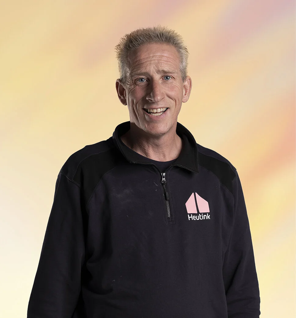 Een glimlachende man met blond haar in een zwarte jas met logo 'Heutink' voor een pastelkleurige achtergrond.