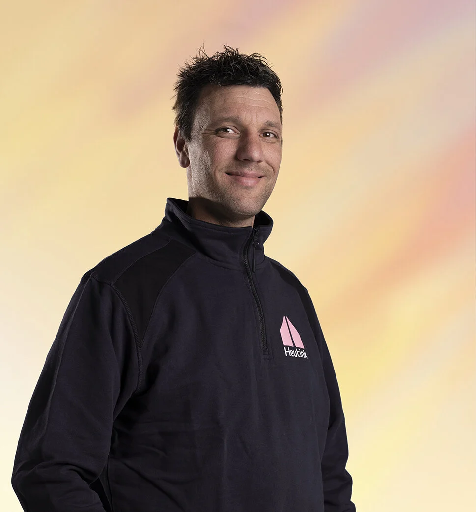 Man in een zwart jack met een logo, glimlachend voor een pastelkleurenachtergrond.