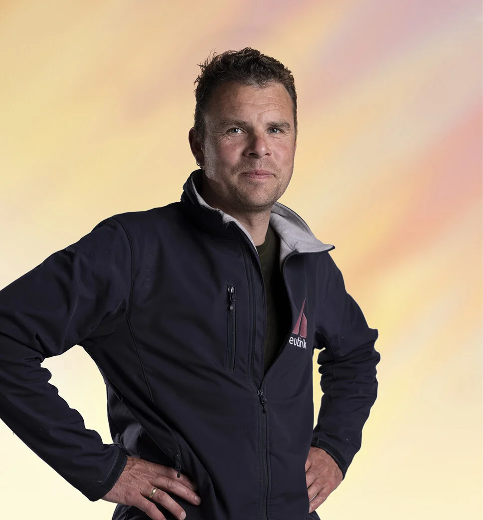 Man staat staand met handen op de heupen, draagt een zwart jasje met een logo op de linker borst, heeft kort bruin haar en kijkt naar de camera, achtergrond is een zacht pastel vierkant met geel en roze tinten.