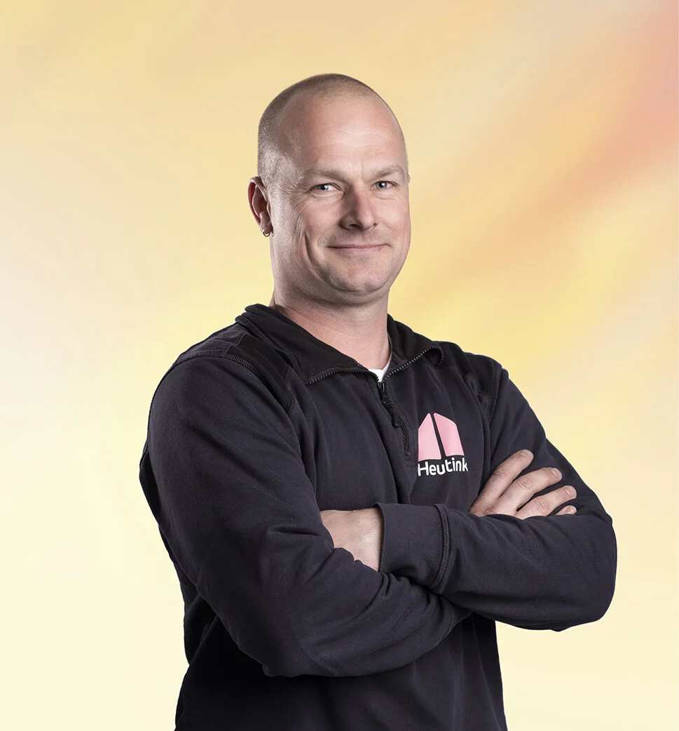 Man met geïsoleerde schedel en donkere jas met logo, achtergrond met zacht gele en roze kleuren.