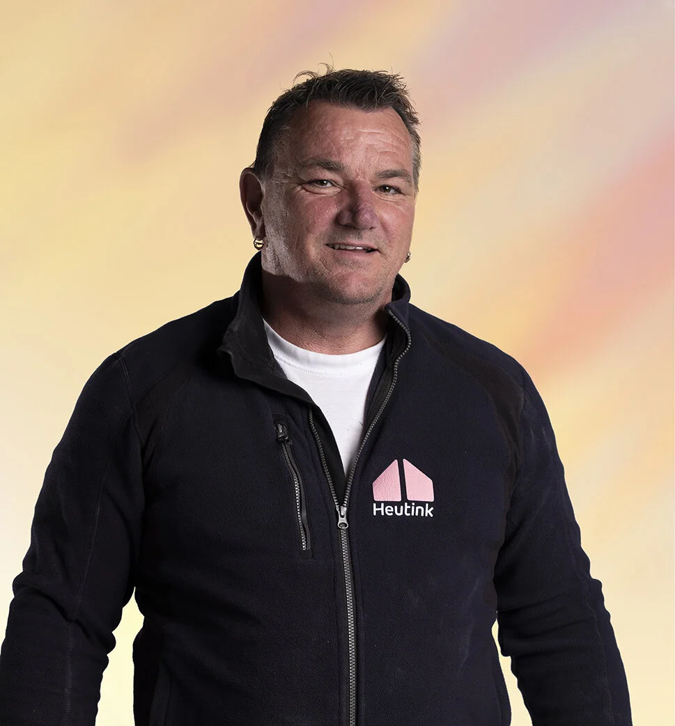 Man met korte haren, oorbellen, in een zwart jack met een logo en tekst 'Heutink' op de borst, staat voor een gekleurde achtergrond.