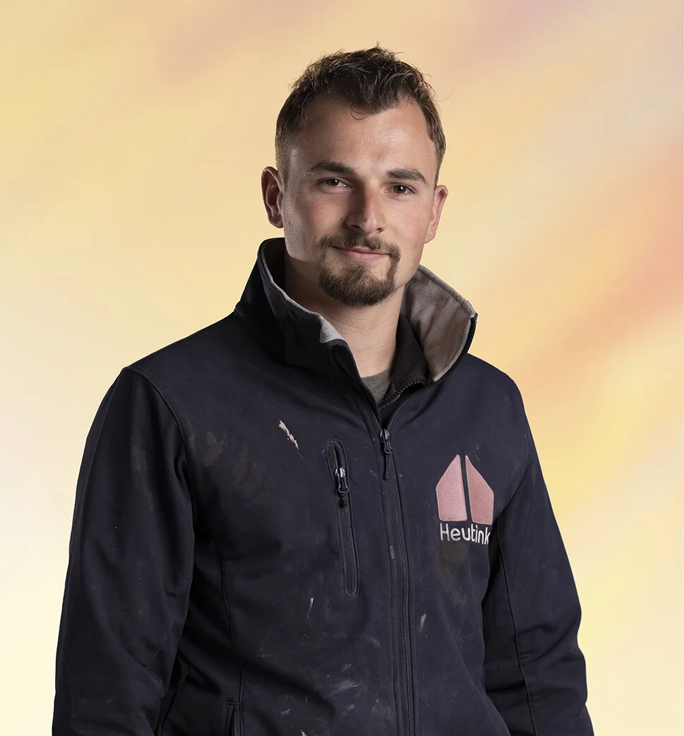 Man met korte haar en een baard, draagt een zwart jack met logo en tekst, op een kleurrijke achtergrond.