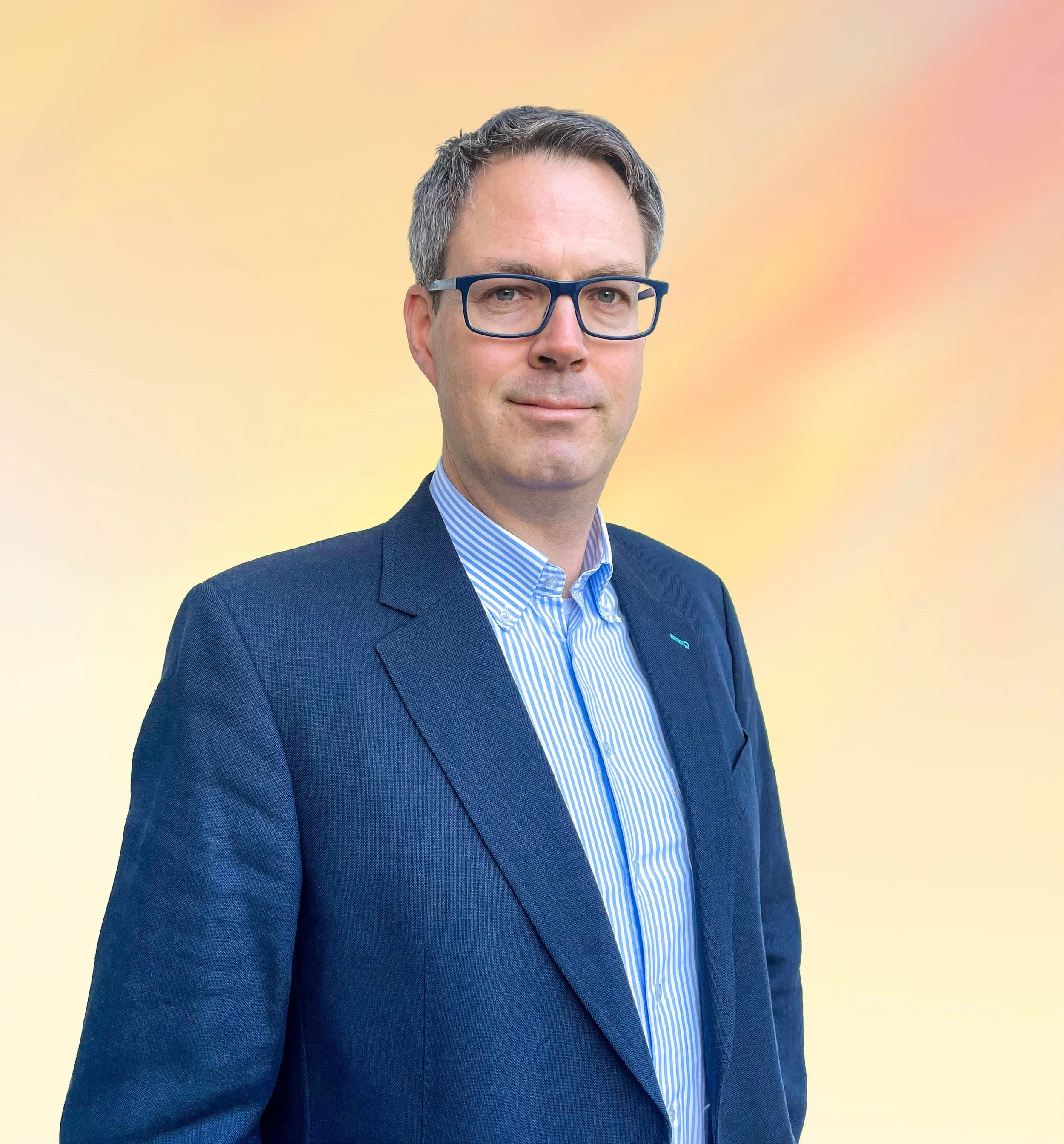 Een man in een donkerblauw kostuum en een blauw-wit gestreept overhemd, draag bril met donkerblauwe montuur, poseert voor een gradiëntachtergrond in pastelkleuren.