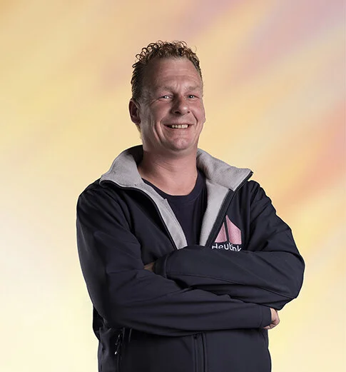 Man met korte, golvende haar die glimlacht en in een zwarte jas staat met zijn armen gekruist, lichte achtergrond met pastelkleuren.