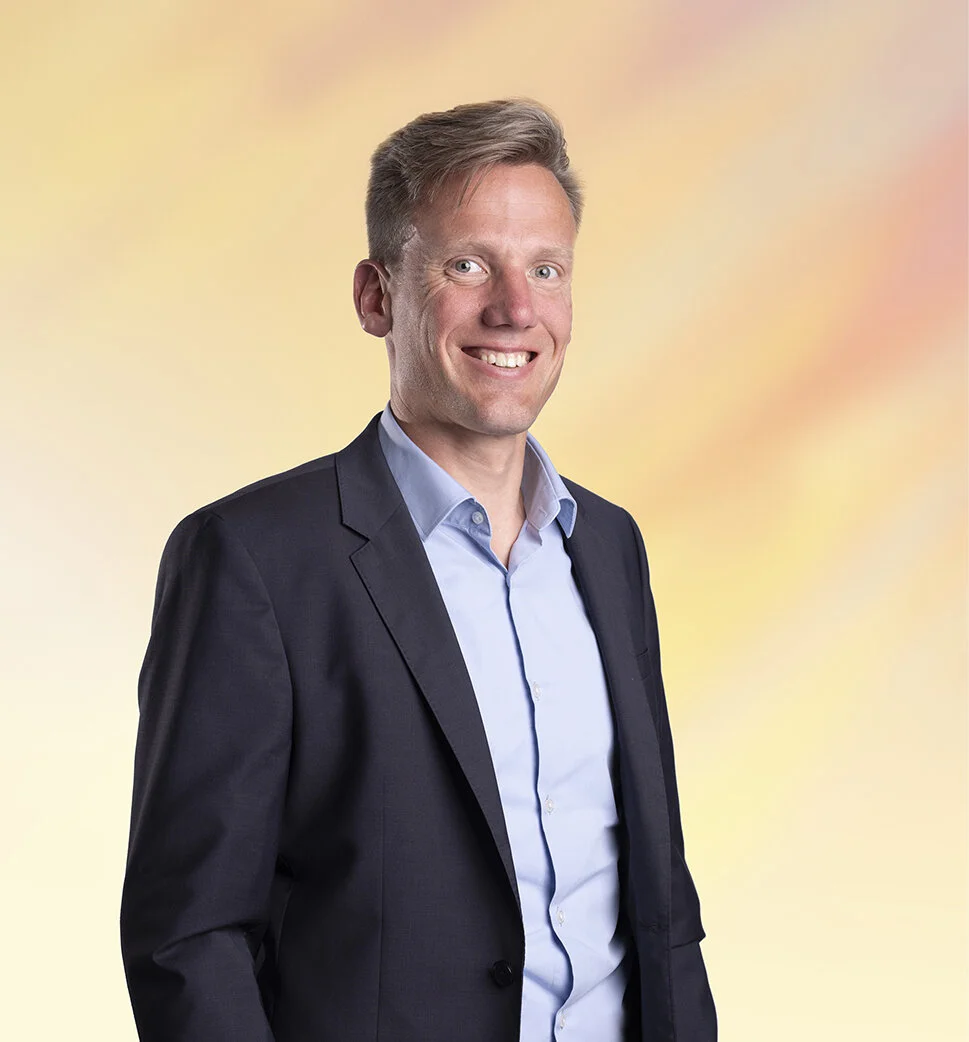 Een lachende man in een zwart pak en een lichtblauwe shirt, poseert voor een kleurrijke, licht vervaagde achtergrond.