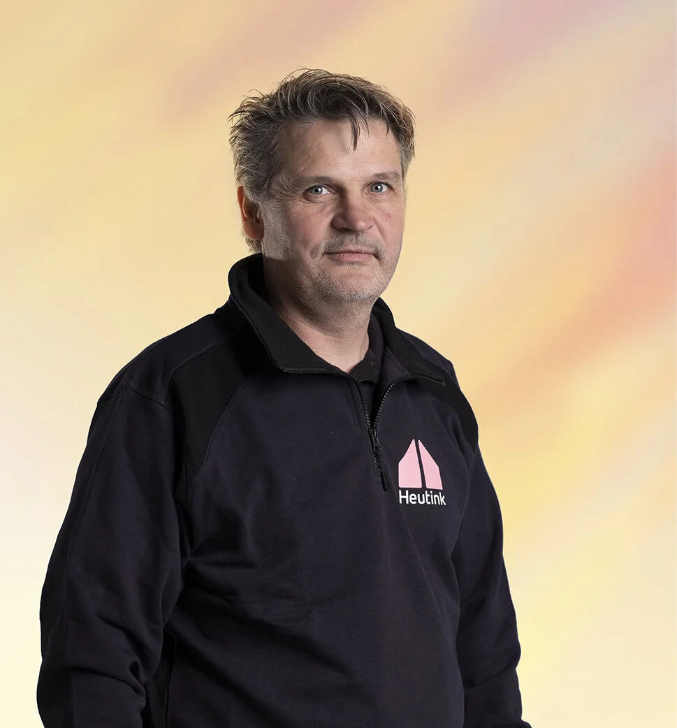 Man in een zwart jasje met een logo en de tekst 'Heutink' op een gekleurde achtergrond.