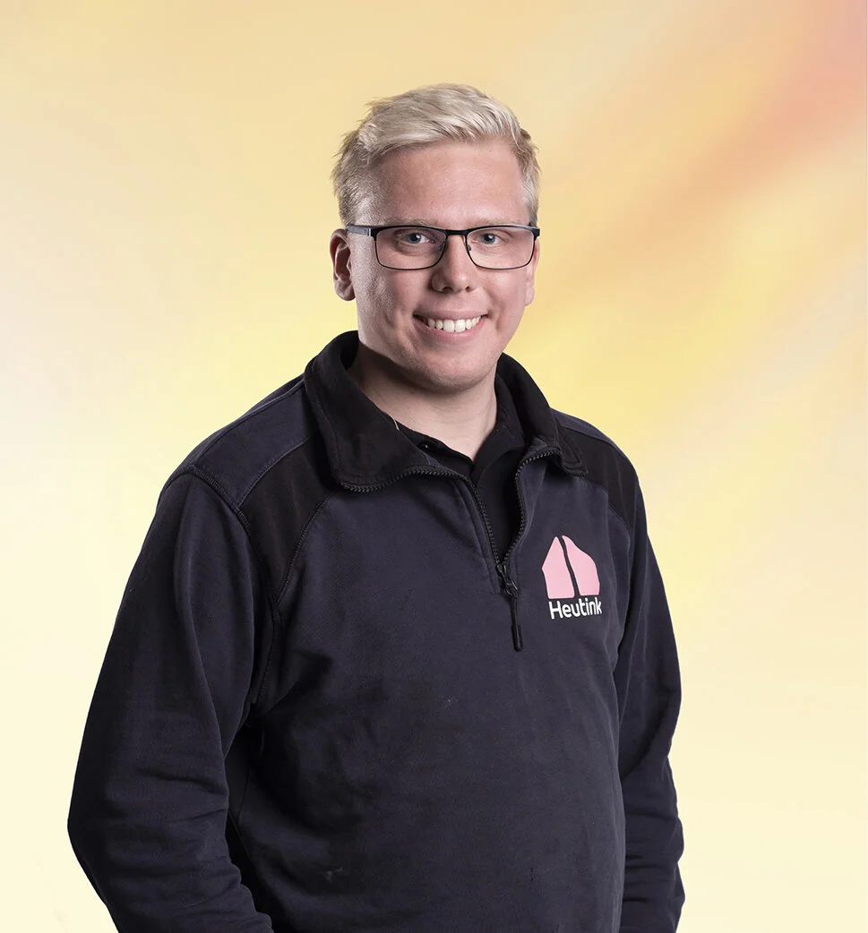 Portret van een lachende man met blond haar, bril, zwarte jas met logo en tekst 'Heutink' voor een pastelkleurige achtergrond.