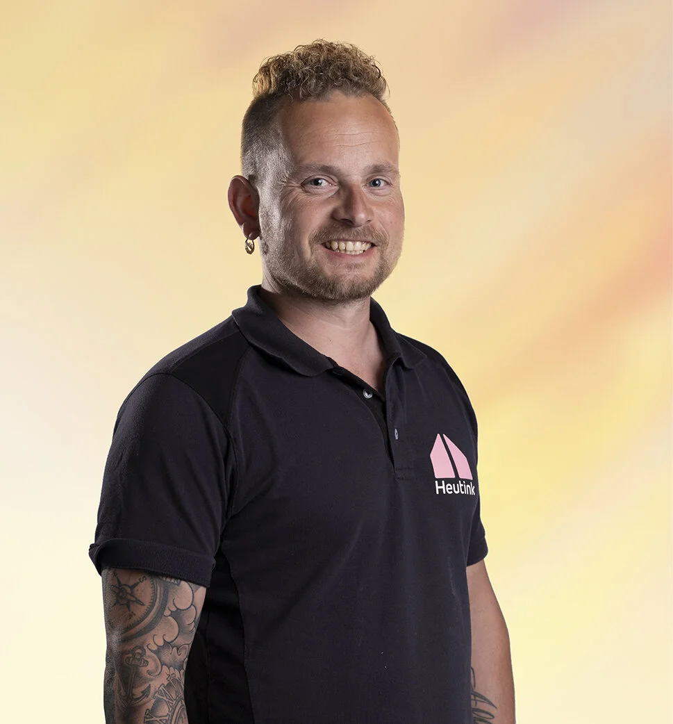 Portret van een man met een glimlach, korte gekrulde haar, een litteken en een tattoo op zijn rechterarm, gekleed in een zwart polo met een logo en tekst, tegen een kleurrijke achtergrond.