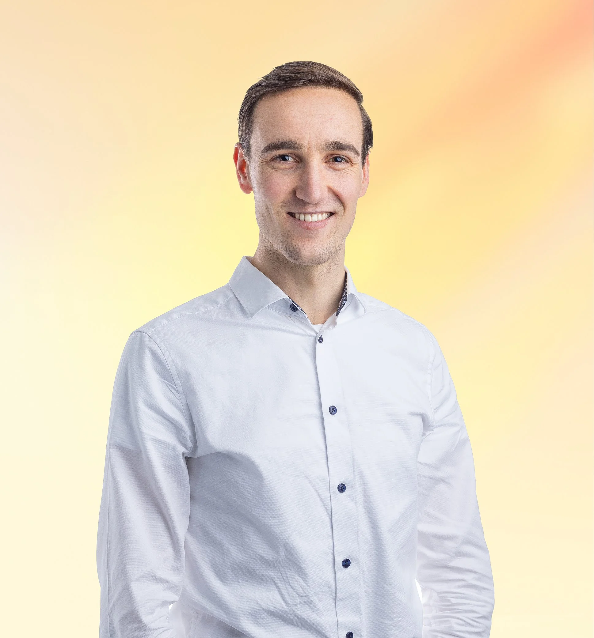 Een jonge man met kort bruin haar draagt een witte blouse en glimlacht, staand voor een zachte gele en oranje achtergrond.