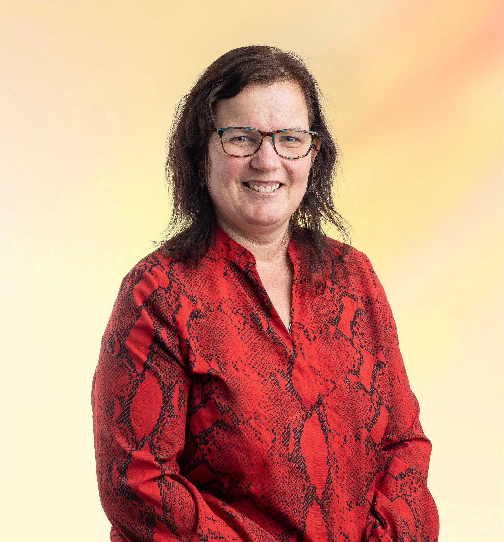 Portret van een vrouw met bruin haar, bril, en een rode blouse met een zwart motief, lachend tegen een geel en roze achtergrond.