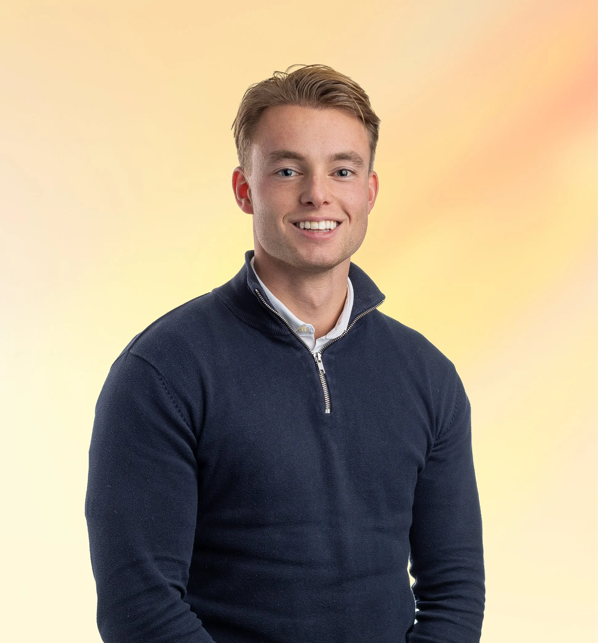 Portret van een jonge man met een lichte haarkleur, glimlach, gekleed in een donkerblauwe trui met rits, witte blouse eronder, voor een zachte gele en roze achtergrond.