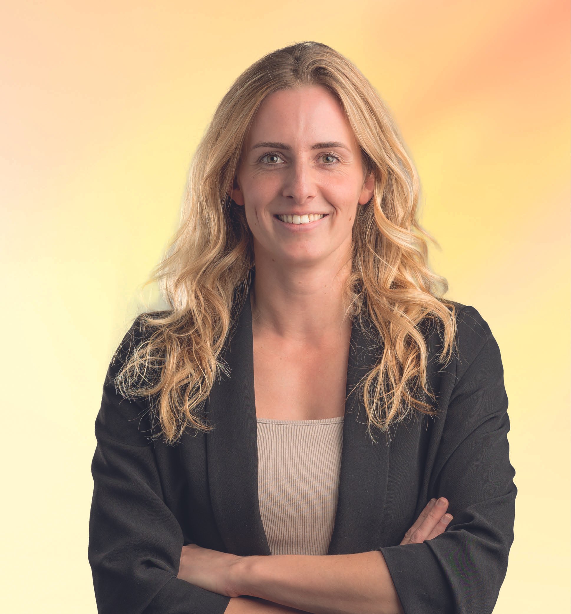 Portret van een lachende vrouw met lang, golvend blond haar, gekleed in een zwart jasje en een beige top, met een gele achtergrond.