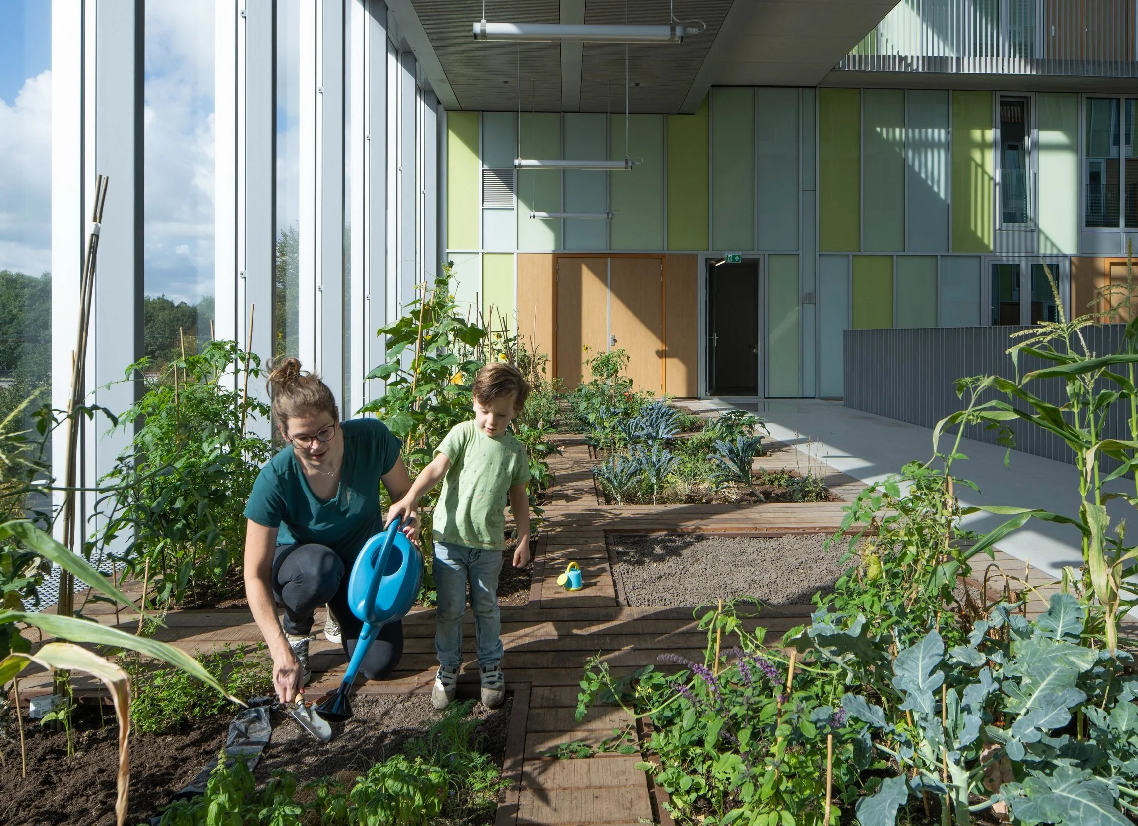 Een vrouw en een jong kind zijn bezig in een binnen-tuin, ze planten en verzorgen groenten en planten, met een moderne, groene gebouw op de achtergrond.