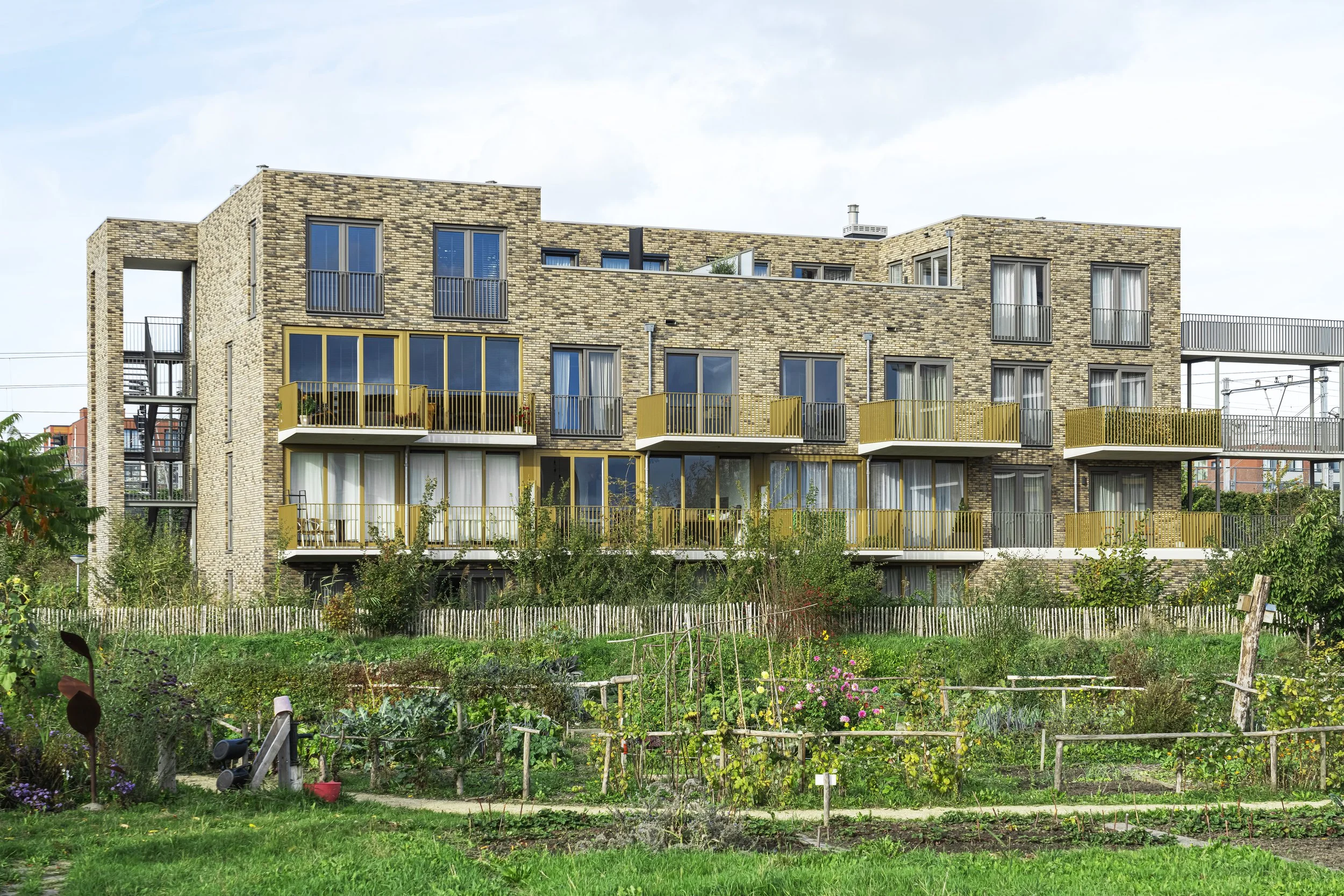 Een modern vier verdiepingen tellend appartementengebouw met grote ramen en balkons, gelegen achter een tuin met moestuin en groen begroeiing.