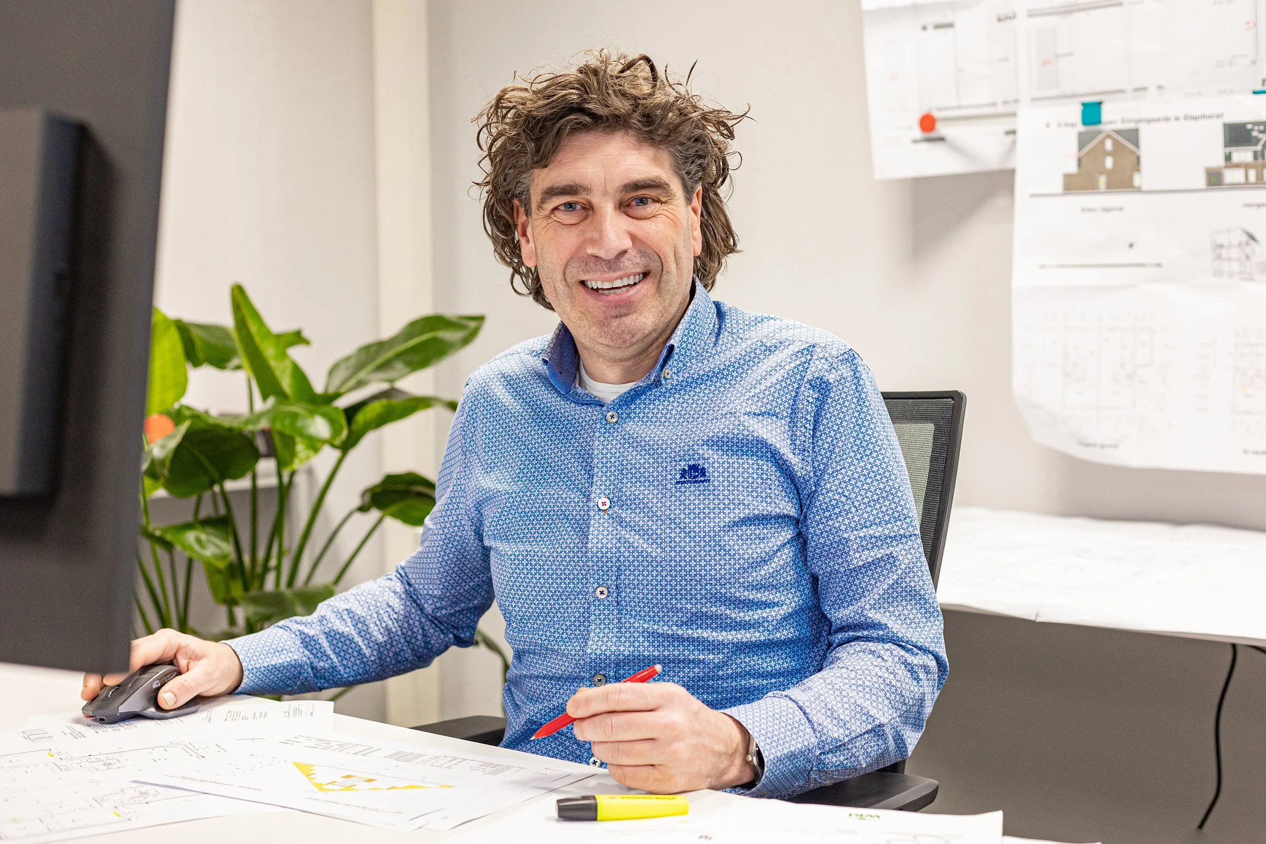 Een man met krullend haar en een blauwe blouse zit aan een bureau met architectuurplannen, een computer en een pen. Er is een groene plant en een schets op de muur in de achtergrond.