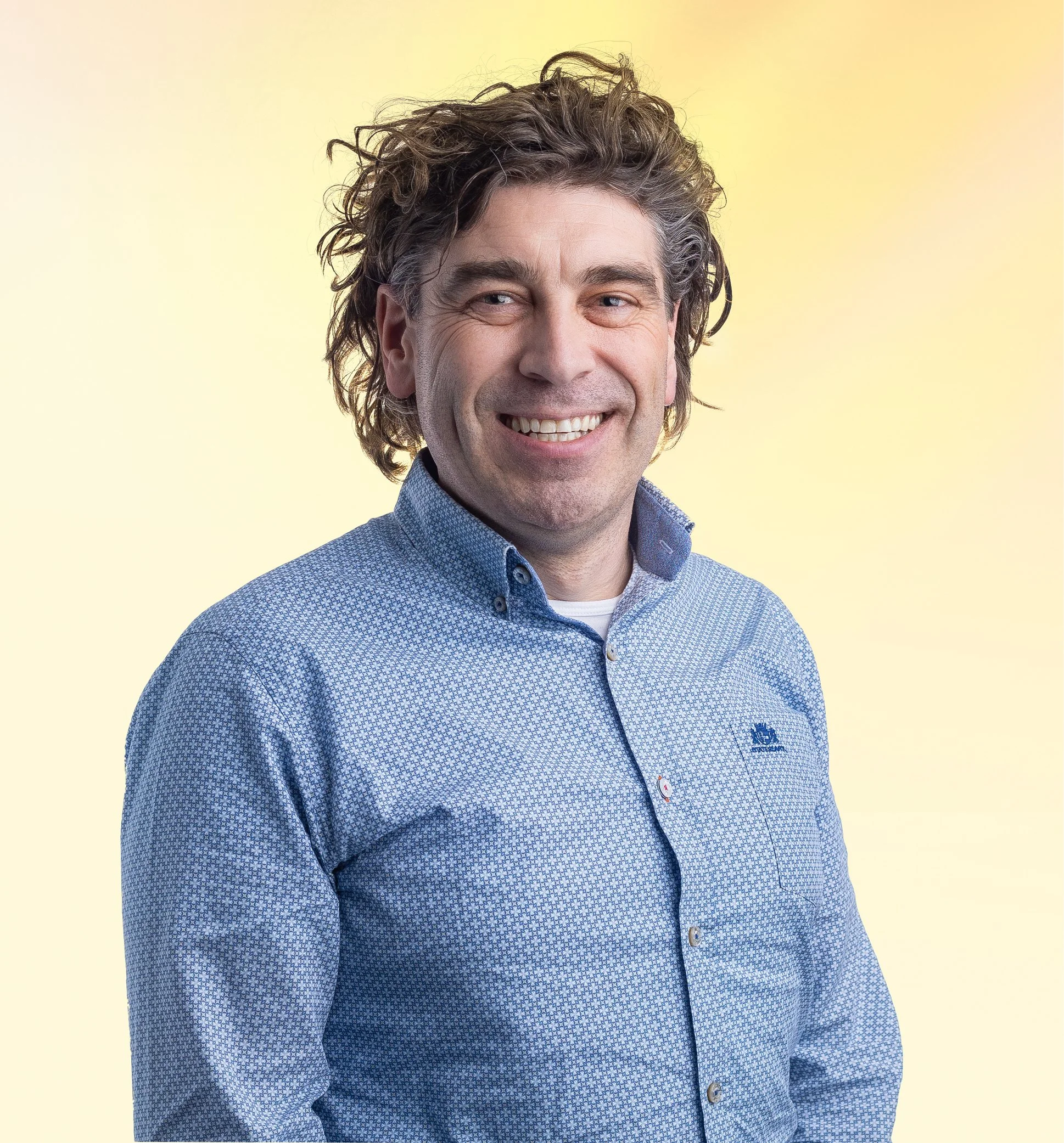 Portret van een lachende man met warrig haar, gekleed in een blauwe, gekleurde blouse, voor een lichtgele achtergrond.