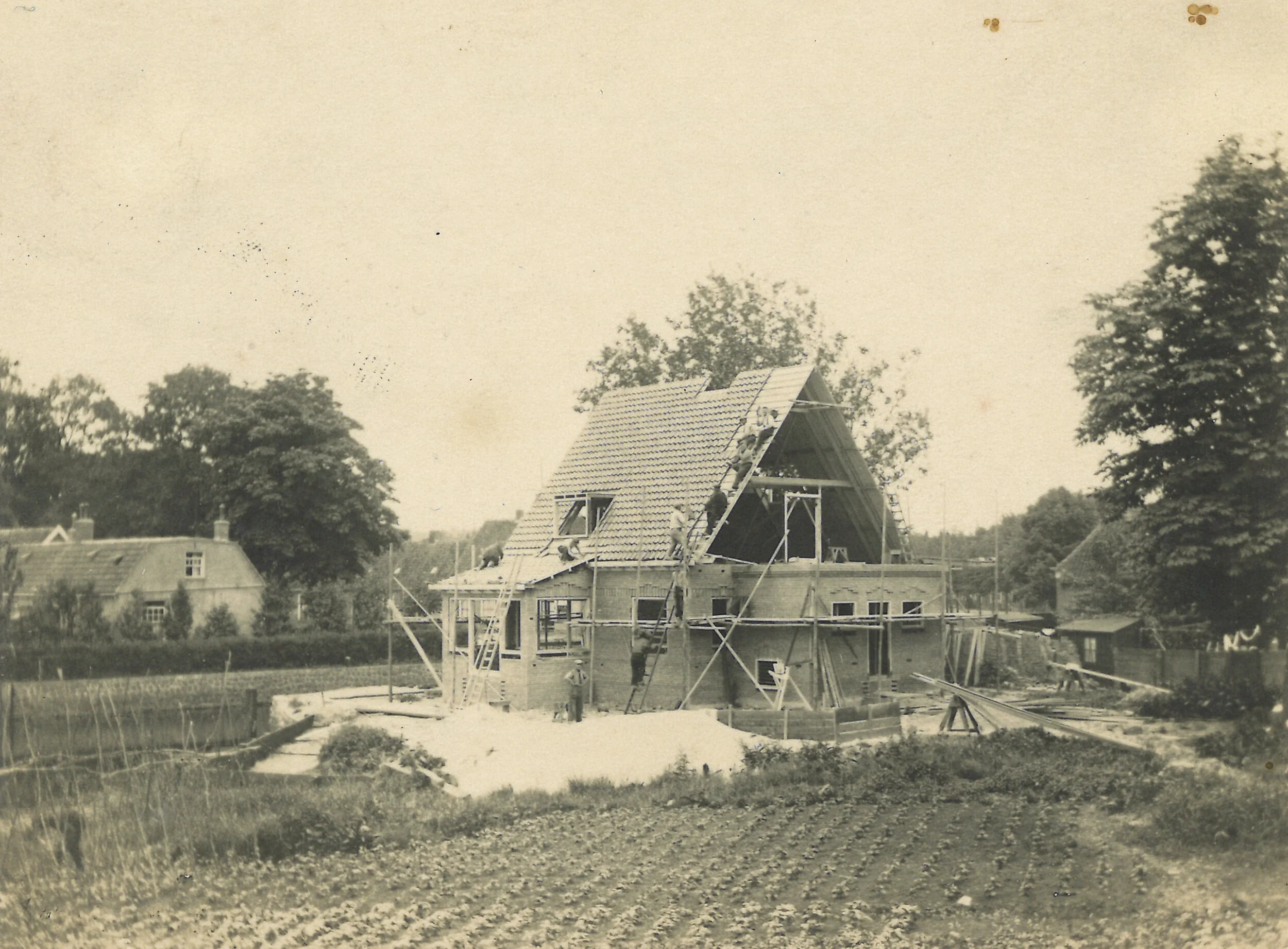 Een zwart-wit foto van een huis dat in aanbouw is, met mensen die op het dak werken en scaffolding eromheen, in een landelijke omgeving.
