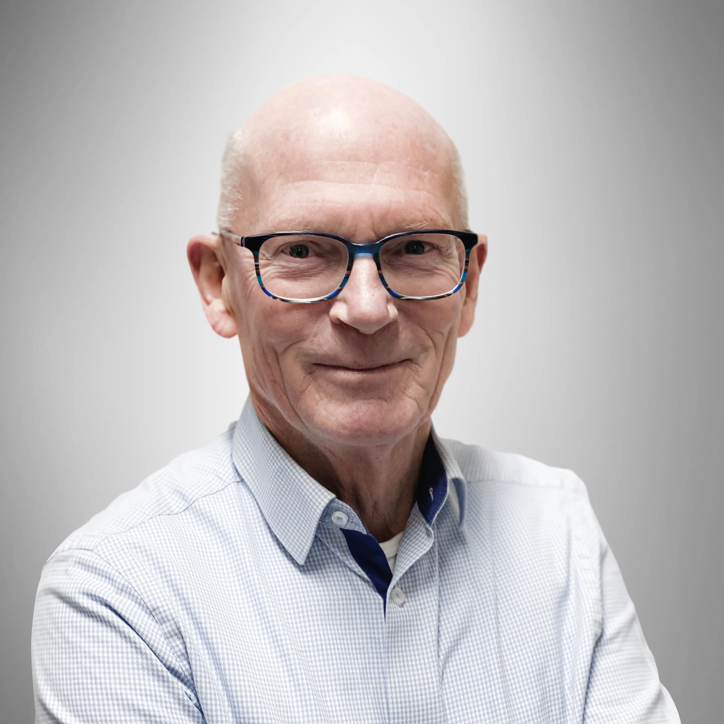 Portret van een oudere man met een glimlach, bril en een lichtblauw geruit overhemd, staand voor een effen lichtgrijze achtergrond.