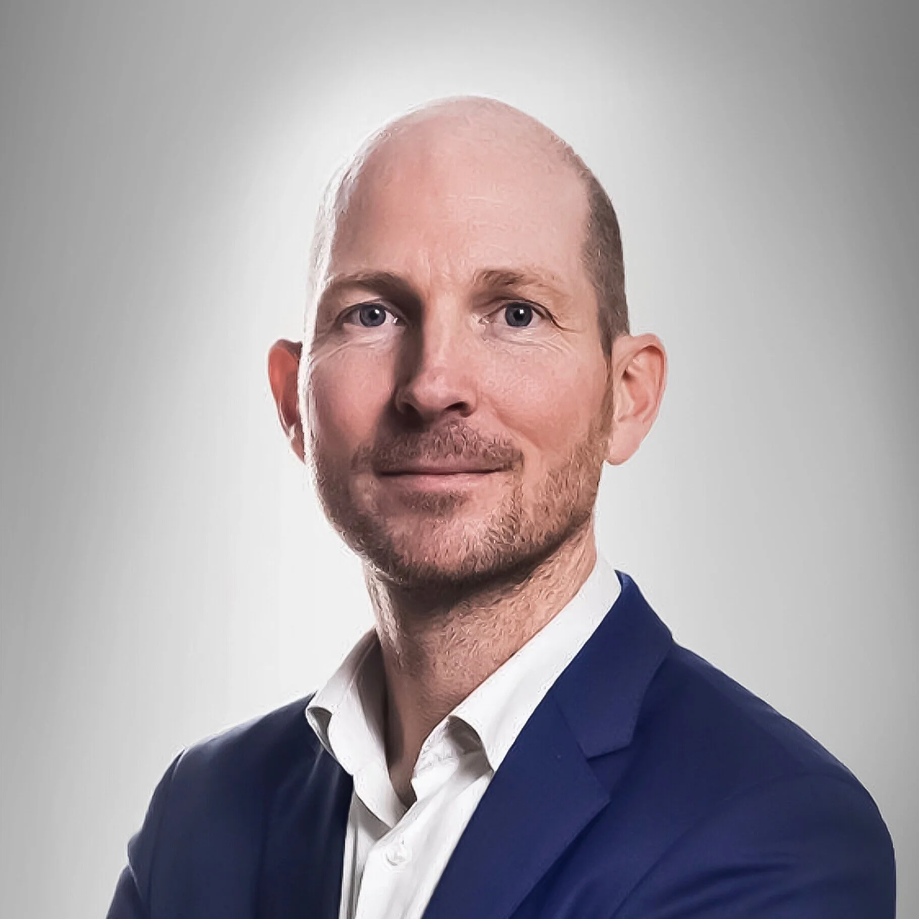 Portret van een man met een blauw pak en een witte overhemd, glimlachend tegen een effen lichte achtergrond.