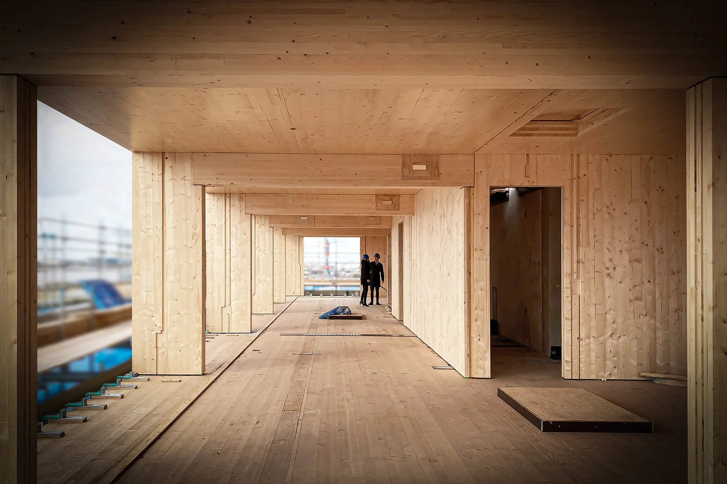 Binnenruimte onder constructie met houten wanden en vloer, twee mensen in de verte met helmen, mogelijk bouwplaats.