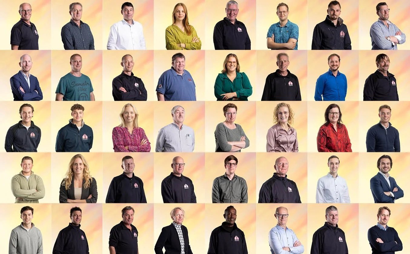 Een groep van 30 mensen, verdeeld over vijf rijen, poseert voor een achtergrond met geel/oranje tinten. De mensen dragen diverse kleding, waaronder zakelijke, casual en sportieve outfits.