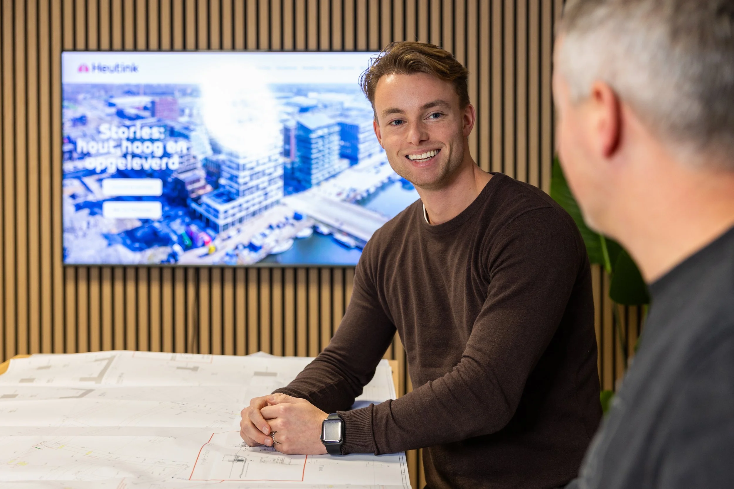 Twee mannen die praten in een vergaderruimte met een grote scherm op de achtergrond en bouwtekeningen op tafel.