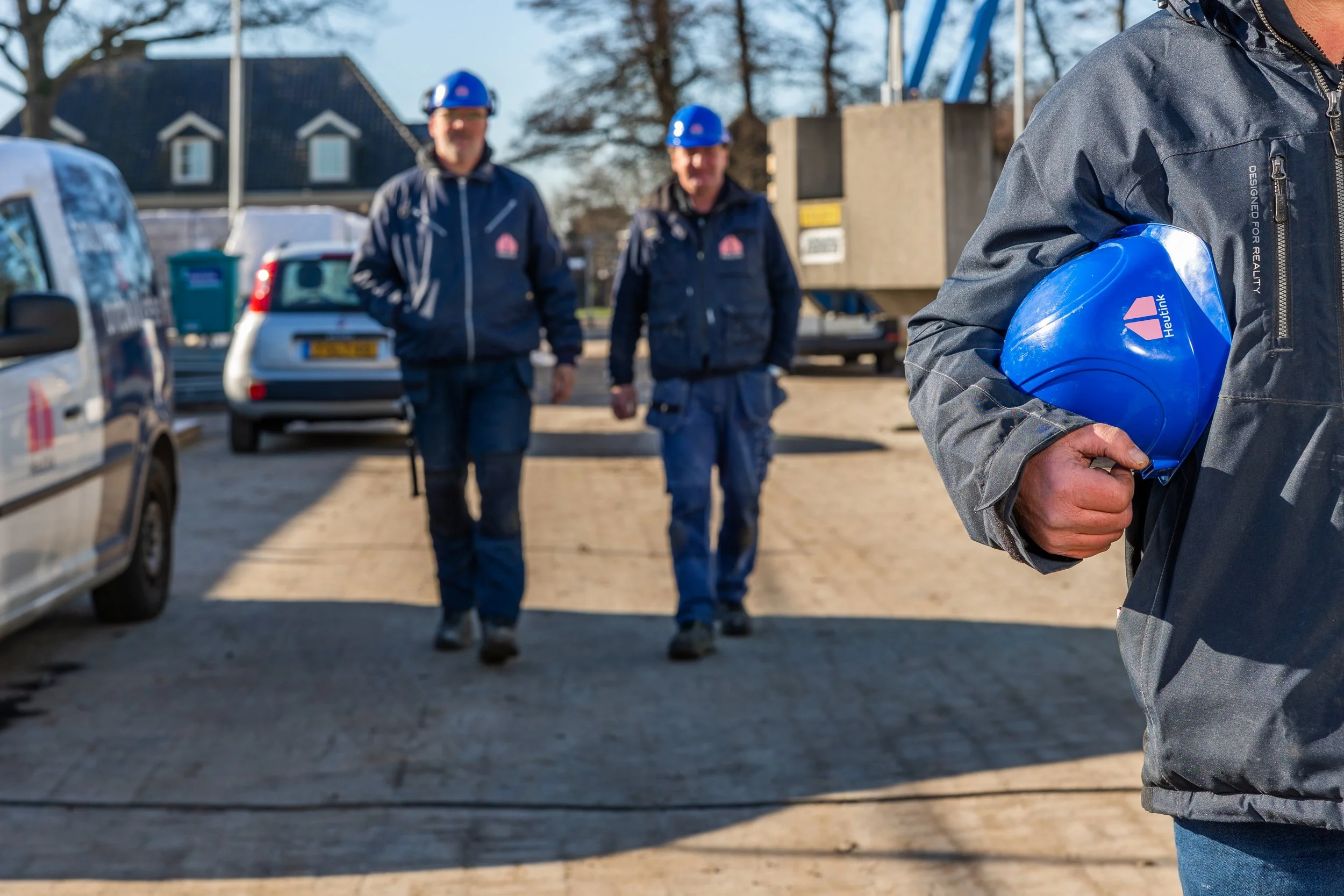 Twee mannen in veiligheidshelmen en jassen lopen op een bouwterrein, terwijl een persoon een blauwe bouwhelm vasthoudt.