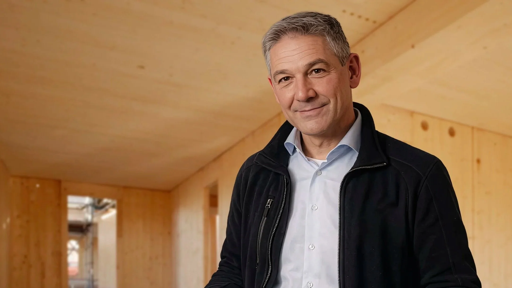 Man in een donkere jas en lichte blouse staat binnen in een houten gebouw, glimlachend naar de camera.